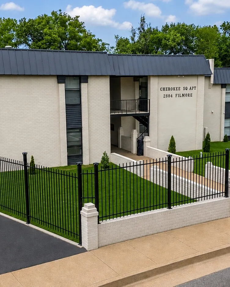 Cherokee Apartments — a cream brick building with a dark metal roof behind a black iron fence and mown lawn