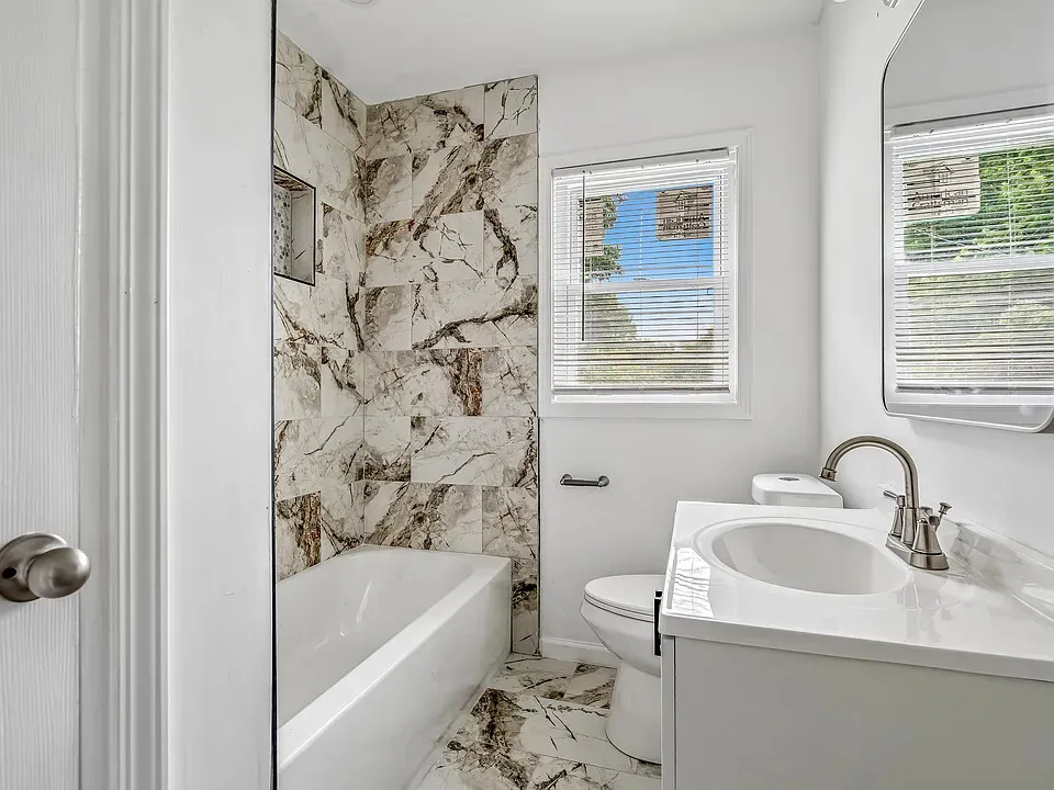 Renovated bathroom with a marble-look tiled tub surround, a bathtub, a blinded window letting in daylight, and a white vanity